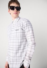 Biela košeľa s dlhým rukávom a čiernym mriežkovým vzorom, s gombíkmi na prednej strane a logom "HUGO" na ľavej hrudi. Model nosí nadrozmerné čierne slnečné okuliare.
