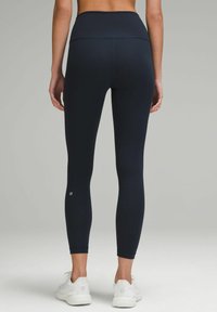 High-waisted, form-fitting leggings in dark navy fabric, featuring a smooth texture and subtle logo at the ankle. Worn with white athletic shoes.