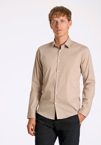 Camicia a maniche lunghe in tessuto beige. Presenta un colletto classico, chiusura con bottoni e bottoni bianchi. Abbinata a pantaloni neri.