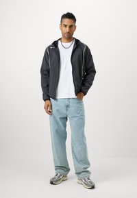 Sort zip-up jakke med hvide accenter, hvid t-shirt, lyseblå jeans og grå sneakers. Model står med en neutral udtryk.