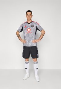 Maglia atletica grigia con motivo geometrico, pantaloni corti neri, calzini bianchi con strisce nere e scarpe bianche, con il logo dello sponsor sul petto.