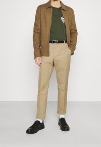 Braune Wollmischung-Jacke mit Reißverschluss über einem grünen T-Shirt, beigen maßgeschneiderten Hosen und schwarzen Sneakers mit weißen Socken. Einfaches Design und Passform.