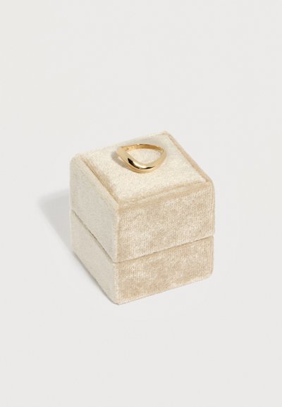 Χρυσό δαχτυλίδι με στριφτό σχέδιο, τοποθετημένο πάνω σε ένα μικρό τετράγωνο βελούδινο κουτί κοσμημάτων σε μπεζ χρώμα, με απλό λευκό φόντο.