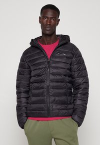 Giacca a vento nera con cappuccio, caratterizzata da un design trapuntato, chiusura con zip frontale e tasche laterali. Indossata sopra una maglietta rossa.