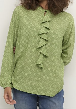 Kobieta nosząca jasnozieloną wzorzystą bluzkę z pionową falbanką na środku i elastycznymi mankietami, w połączeniu z niebieskimi dżinsami.