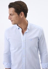 Camisa de botón a rayas azul claro y blanca, hecha de una tela ligera. Presenta un sutil logo bordado y mangas enrolladas.