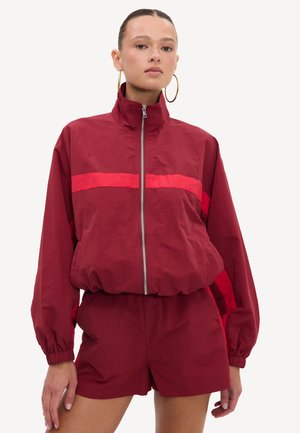 Femme portant une veste bordeau zippée avec des rayures rouges et un short assorti, debout, les mains détendues le long du corps, sur fond blanc.