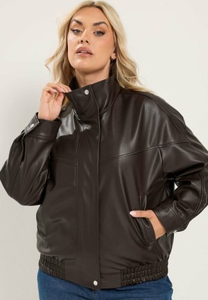 Femme aux cheveux blonds portant une veste en cuir marron foncé avec un col haut, une main ajuste le col, l’autre main dans la poche.