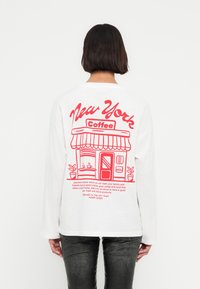 Λευκό πουκάμισο με μακριά μανίκια που διαθέτει μια κόκκινη εικόνα από καφετέρια και κείμενο, με στρογγυλή λαιμόκοψη και άνετη εφαρμογή.