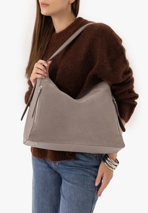 Woman wearing brown sweater and blue jeans holding a large gray shoulder bag with zipper details and silver bracelets on her wrist.