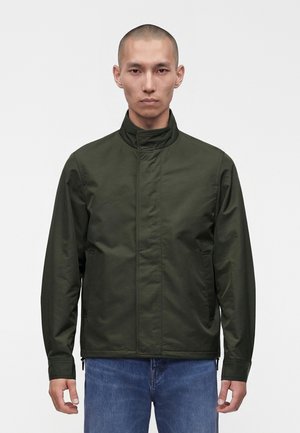 Homme aux cheveux courts portant une veste zippée vert foncé et un jean bleu, debout devant un fond blanc uni.
