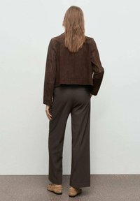 Giacca cropped in camoscio marrone con una texture liscia, abbinata a pantaloni ampi scuri. L'outfit presenta un design minimalista e colori neutri.