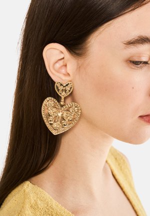 Orecchini a forma di cuore in tonalità oro con intricati motivi floreali, dotati di un accento a forma di cuore più piccolo sopra. La superficie testurizzata esalta i dettagli.