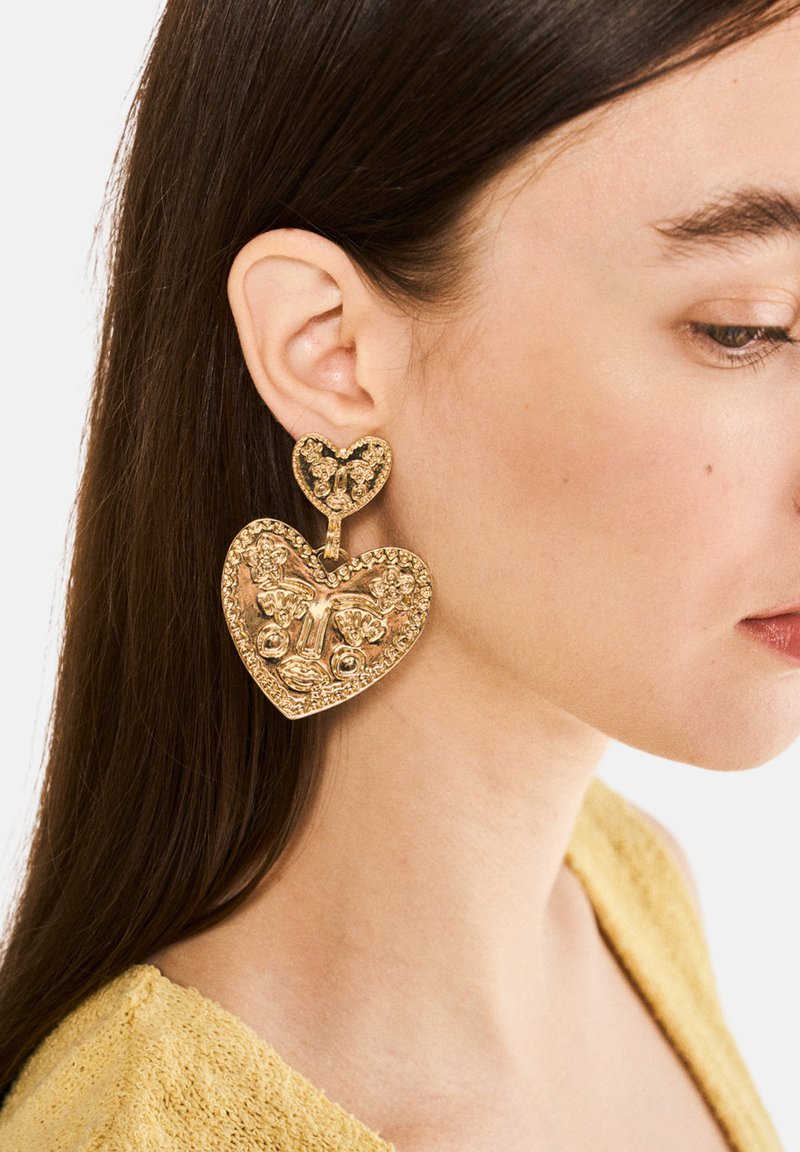 Pendientes en forma de corazón tono dorado con intrincados patrones florales, que presentan un pequeño corazón como acento en la parte superior. La superficie texturizada realza los detalles.