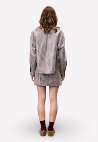 Persoon met lang golvend haar, gekleed in een geruit shirt en bijpassende rok, donkere sokken en pluizige pantoffels, staand met de rug naar de camera tegen een witte achtergrond.