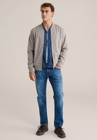 Hall hallit peakoontega bomberi jope, mille all sinine nööbitav särk, kombineeritud helesinine teksade ja pruunide pits-up kingadega.