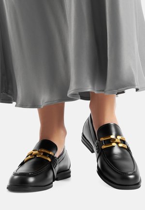 Schwarze Leder-Loafers mit glänzendem Finish, verziert mit einer goldfarbenen Schnalle und kleinem Blockabsatz. Kombiniert mit einem grauen, fließenden Rock.