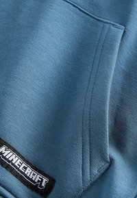 Blauer Stoff-Hoodie mit strukturierter Oberfläche, einer Fronttasche und einem schwarzen "Minecraft"-Logopatch.
