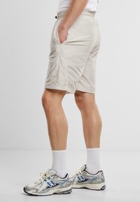 Beige Sportshorts mit einem elastischen Bund, seitlichen Taschen und weißen Paspelierungen. Getragen mit grauen und blauen Turnschuhen und weißen Socken.