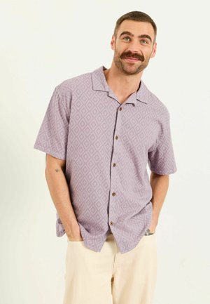 Mann mit Schnurrbart, der ein gemustertes kurzärmliges Hemd mit Knöpfen und helle Hosen trägt, mit Händen in den Taschen stehend und lächelnd.