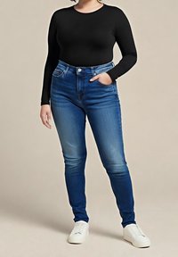 Jean skinny bleu taille haute avec un lavage délavé, poches avant et passants de ceinture. Associé à un haut noir à manches longues et des baskets blanches.