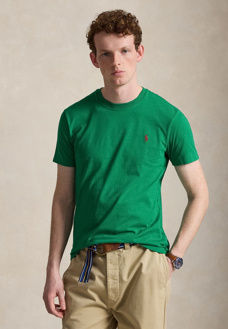 Ung mand med krøllet hår iført grøn kortærmet skjorte, beige bukser med stribet bælte og et armbåndsur, stående mod en ensfarvet baggrund.