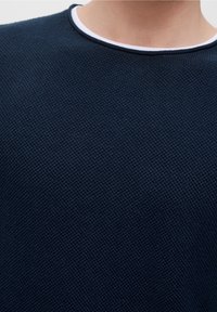 Lähikuva henkilöstä, joka käyttää tummansinistä, teksturoitua neuletta, jossa on valkoinen ja tummansininen pyöreä kaulus.