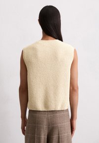 Cremfarbener, strukturierter, ärmelloser Pullover mit geripptem Muster, geradem Schnitt und rundem Hals; kombiniert mit braunen karierter Hose.