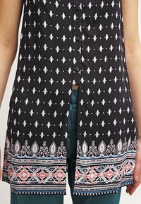 Czarna tunika zapinana na guziki z białymi geometrycznymi wzorami, rozcięciem z przodu oraz ozdobnym dołem z kolorowymi kwiatowymi i plemiennymi motywami.