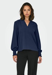 Femme aux cheveux mi-longs portant un chemisier bleu marine à manches longues avec des poignets froncés et un pantalon noir, debout devant un fond uni.