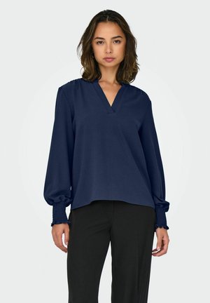 Femme aux cheveux mi-longs portant un chemisier bleu marine à manches longues avec des poignets froncés et un pantalon noir, debout devant un fond uni.