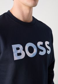 Jūras spēku džemperis ar apaļu kakla izgriezumu, uz kura ir liels "BOSS" logo gaiši zilā krāsā, izgatavots no gludas, teksturētas auduma.