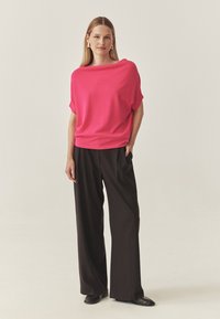 Rosa, oversized topp med bred halsringning som matchas med svarta vida byxor. Modell som bär texturerade svarta platta skor. Neutral bakgrund.