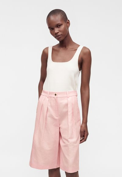 Femme aux cheveux courts portant un haut blanc sans manches et un short large rose taille haute, debout devant un fond uni.