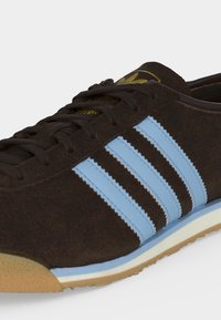 Σκούρο καστανό sneaker από σουέντ με τρεις ανοιχτό μπλε ρίγες στη πλευρά, ανοιχτό μπλε και λευκή σόλα και λαστιχένια προστασία μύτης.