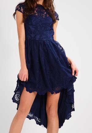 Robe bleu marine en dentelle à manches courtes, corsage ajusté et ourlet festonné haut-bas portée par une femme debout.