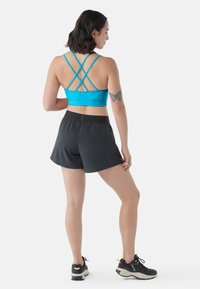 Sostén deportivo azul con tirantes cruzados, combinado con pantalones cortos negros con cinturilla elástica. La modelo lleva zapatillas deportivas negras.