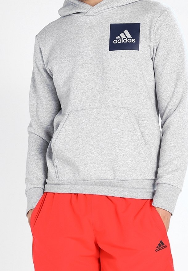 Personne portant un sweat à capuche Adidas gris clair avec le logo sur la poitrine et un short Adidas rouge vif avec le logo sur la cuisse gauche, les mains dans les poches.