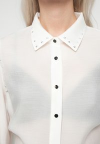 Balta marškinėliai su permatomu tekstūra, su apykakle, papuošta sidabro kaiščiais ir juodais mygtukais priekyje.