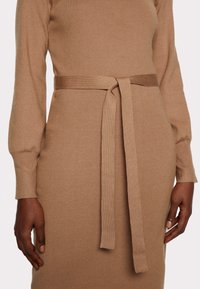 Lähellä otettu kuva henkilöstä, joka käyttää mukautettua, pitkähih lasta beigeä neulemekkoa, jossa on sidottu vyö vyötäröllä.