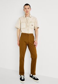 Short-sleeved beige shirt with two front pockets, paired with brown trousers and black sneakers featuring white soles and laces.