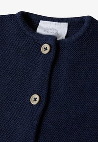 Marineblaue Strickjacke mit rundem Kragen, ausgestattet mit zwei Holzknöpfen und einem weißen Stoffetikett mit der Aufschrift "Stellou & Friends."