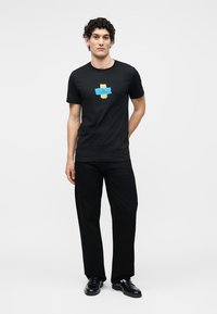 Jeune homme debout, les mains dans le dos, portant un t-shirt graphique noir, un pantalon noir et des chaussures habillées noires sur un fond uni.