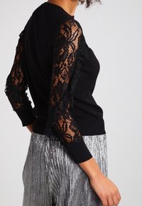 Černý svetr s průhlednými květinovými krajkovými dlouhými rukávy v kombinaci s metalickým stříbrným skládacím sukní na osobě před jednobarevným pozadím.