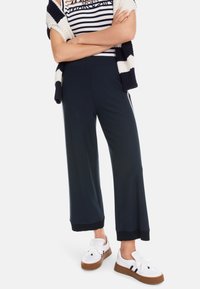 Weite marineblaue Hose mit einer glatten Textur, ausgestattet mit einem kontrastierenden schwarzen Saum. Kombiniert mit weißen Sneakern und einem gestreiften Oberteil.