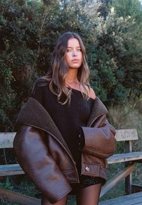 Överdimensionerad brun läderjacka med ett fårskinnskrage, svart ribbad off-shoulder tröja och svarta paljettshorts, mot en grönskande bakgrund.