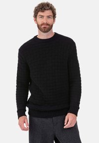 Schwarzer Strickpullover mit strukturierter Webart, rundem Ausschnitt sowie gerippten Bündchen und Saum. Kombiniert mit grauer Hose.