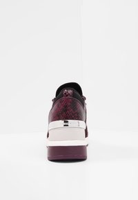 Baksidan av en lila sneaker med snakeskin-textur, silverdetalj och tjock sula på en vit yta och bakgrund.