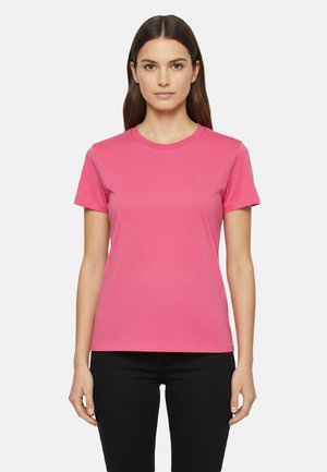 Donna con capelli scuri lisci, indossa una maglietta rosa semplice a maniche corte e pantaloni neri, in piedi davanti a uno sfondo bianco.