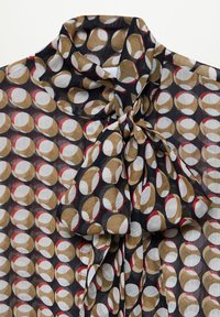 Blusa transparente con fondo negro y un patrón circular en dorado, blanco y rojo. Cuello atado con tela drapeada para textura.